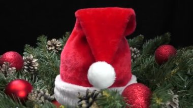Kırmızı Noel Baba şapkası yeşil kozalak dalları ve noel toplarıyla çevrili.