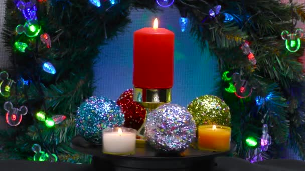 在圣诞花圈和照明的背景下 一个由新花球和小蜡烛环绕的燃烧的蜡烛旋转着 — 图库视频影像
