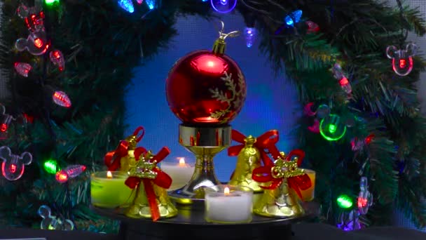 鐘と小さなろうそくに囲まれた赤いクリスマスボールは クリスマスの花輪と照明の背景に回転します — ストック動画