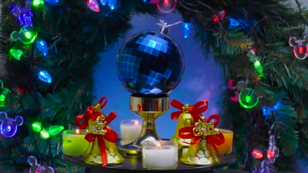 在圣诞花圈和照明的背景下 一个蓝色格子的圣诞球被铃铛和小蜡烛环绕着旋转着 — 图库视频影像