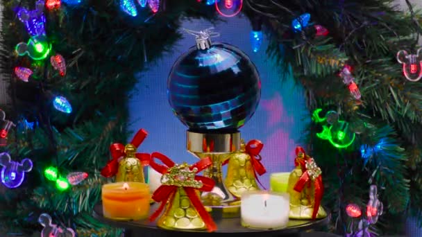 一个蓝色格子的圣诞球 四周环绕着铃铛和小蜡烛 在明亮的圣诞花环和照明的背景下旋转着 — 图库视频影像