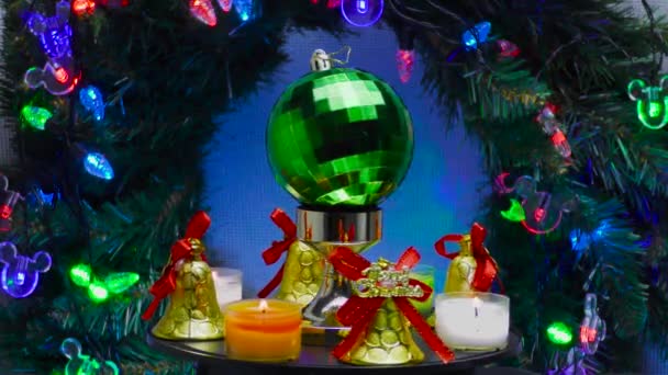 鐘と小さなろうそくに囲まれた緑の格子状のクリスマスボールは クリスマスの花輪と照明の背景に回転します — ストック動画