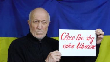 Ukrayna bayrağının arka planında duran yaşlı bir adam Ukrayna üzerinde gökyüzünü kapatan bir işaret kaldırıyor.