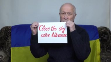 Ukrayna bayrağının arkasına yaslanmış yaşlı bir adam Ukrayna semalarında bir işaret kaldırıyor.
