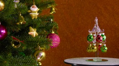 On a colored background, a small Christmas tree rotates near a beautiful decorated Christmas tree
