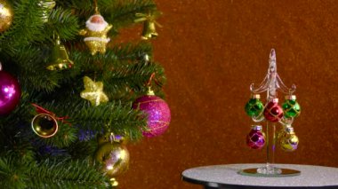 On a colored background, a small Christmas tree rotates near a beautiful decorated Christmas tree