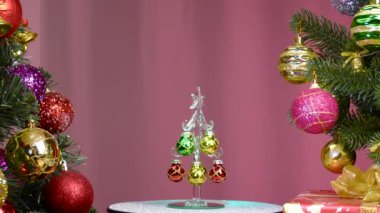 On a pink background, a small Christmas tree rotates near large Christmas trees decorated with Christmas decorations