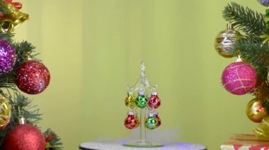 On a yellow background, a small Christmas tree rotates near large Christmas trees decorated with Christmas decorations
