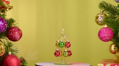 On a yellow background, a small Christmas tree rotates near large Christmas trees decorated with Christmas decorations
