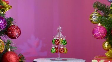 On a pink background, a small Christmas tree rotates near large Christmas trees decorated with Christmas decorations