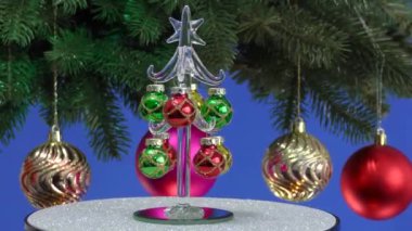 Mavi arka planda, küçük bir Noel ağacı Noel toplarıyla süslenmiş dönen büyük bir Noel ağacının yanında döner.