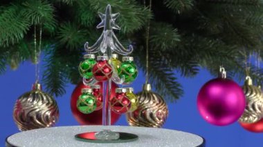 Mavi arka planda, küçük bir Noel ağacı Noel toplarıyla süslenmiş dönen büyük bir Noel ağacının yanında döner.