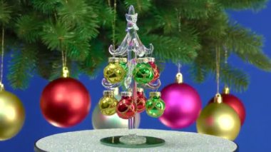 Mavi arka planda, küçük bir Noel ağacı Noel toplarıyla süslenmiş dönen büyük bir Noel ağacının yanında döner.