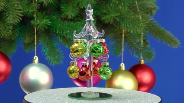 青い背景に小さなクリスマスツリーがクリスマスボールで飾られた回転する大きなクリスマスツリーの近くに回転します — ストック動画