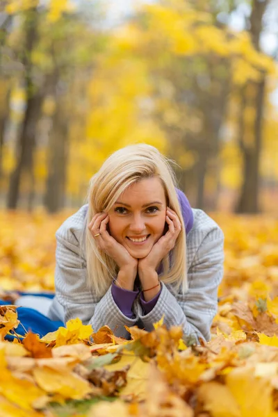 Portrait of a joyful young woman laying on dry leaves in autumn park. Beautiful blonde female in autumn light grey coat and blue hoodie. Relax in nature.