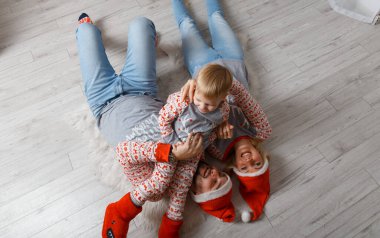 Noel ağacının yanında mutlu bir aile yerde eğleniyor. Evde Noel kutlaması. Anne, baba ve oğul rahat giyindi. Mutlu aile eğlencesi yatakta oynuyor. Noel süslemeli gri iç döşeme