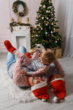 Noel ağacının yanında mutlu bir aile yerde eğleniyor. Evde Noel kutlaması. Anne, baba ve oğul rahat giyindi. Mutlu aile eğlencesi yatakta oynuyor. Noel süslemeli gri iç döşeme