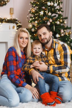 Noel ağacının yanında mutlu bir aile yerde eğleniyor. Evde Noel kutlaması. Anne, baba ve oğul rahat giyindi. Mutlu aile eğlencesi yatakta oynuyor. Noel süslemeli gri iç döşeme