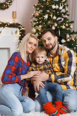 Noel ağacının yanında mutlu bir aile yerde eğleniyor. Evde Noel kutlaması. Anne, baba ve oğul rahat giyindi. Mutlu aile eğlencesi yatakta oynuyor. Noel süslemeli gri iç döşeme