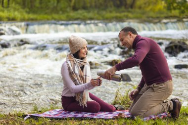 Genç ve güzel bir çift sonbahar nehrinin kıyısında dinleniyor ve kendini termostan gelen sıcak bir içecekle ısıtıyor..