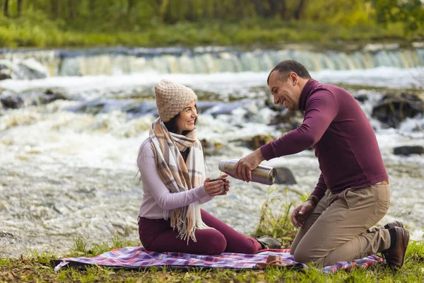 Genç ve güzel bir çift sonbahar nehrinin kıyısında dinleniyor ve kendini termostan gelen sıcak bir içecekle ısıtıyor..