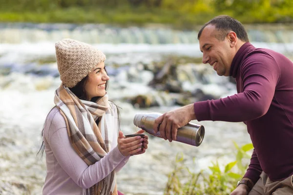 Genç ve güzel bir çift sonbahar nehrinin kıyısında dinleniyor ve kendini termostan gelen sıcak bir içecekle ısıtıyor..