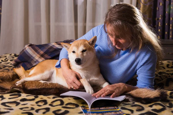 ベッドの上に寝そべって仲間の柴犬と本を読んでいる女性 愛と団結とペットを屋内で — ストック写真