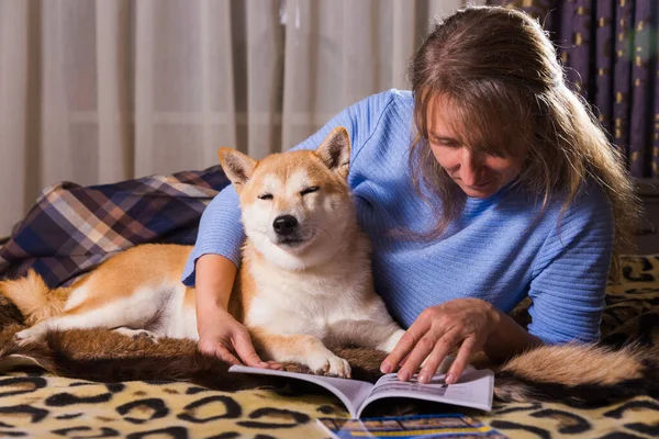 ベッドの上に寝そべって仲間の柴犬と本を読んでいる女性 愛と団結とペットを屋内で — ストック写真