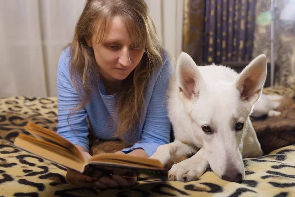 ベッドに横になり 仲間の白いスイスの羊飼いの犬と本を読んでいる女性 愛と団結とペットを屋内で — ストック写真