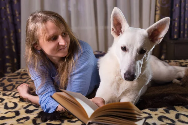 ベッドに横になり 仲間の白いスイスの羊飼いの犬と本を読んでいる女性 愛と団結とペットを屋内で — ストック写真