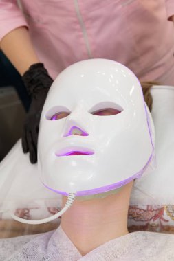 Kadın kurşun terapi maskesiyle yatakta uzanıyor ve rahatlıyor. Yüz bakımı için öncü hafif maske, dondurucu kozmetoloji prosedürü.