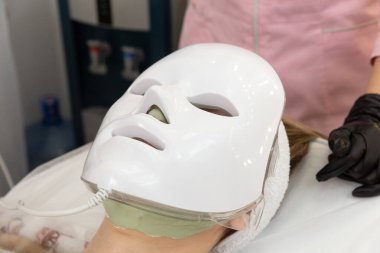 Kadın kurşun terapi maskesiyle yatakta uzanıyor ve rahatlıyor. Yüz bakımı için öncü hafif maske, dondurucu kozmetoloji prosedürü.