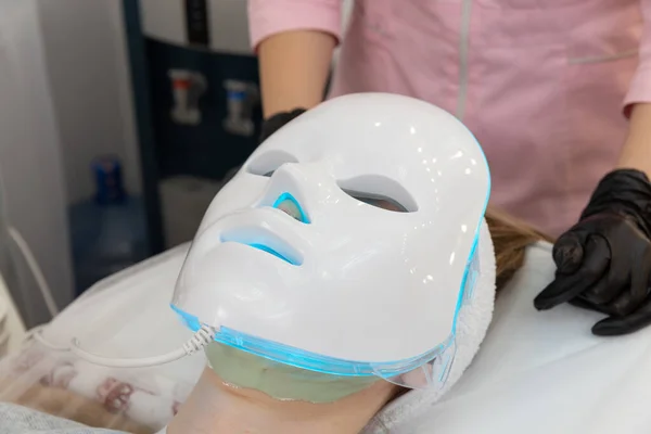 Woman lies in bed with led light therapy facial mask and relax. Led light mask for face skin treatment, frofessional cosmetology procedure.