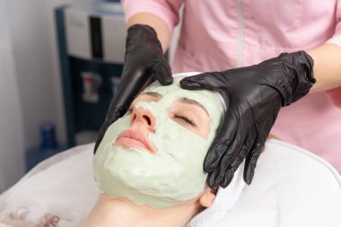 Lastik eldivenli bir kozmetolog müşteriye krem maske takar ve yüz masajı yapar..