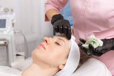 Güzellik uzmanı, kaplıca salonundaki genç bir kadının yüzünden kilden bir maske çıkarıyor. Güzellik uzmanındaki güzel genç kız spa prosedürlerini yapıyor.