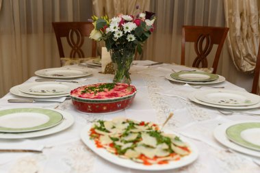 Tabaklar ve sofra takımları şık ev dekorasyonunda. İnsan yok. Yer ayarlı yemek masası. Örgü peçeteler, kadehler ve çiçekli vazolar.