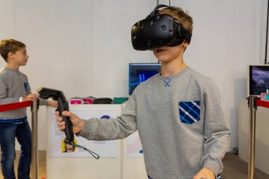 Sekiz yaşındaki çocuk VR kulaklık takıyor, elinde kontrolörlerle video oyununda yeni deneyimlerin tadını çıkarıyor. Modern teknolojiler sergisinde oyun standı.
