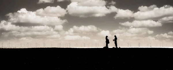 2 people on a shore line with off shore wind turbines in the distance in mono chrome