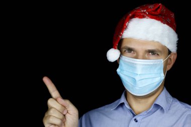 Noel kutlaması, maskeli adam ve Noel Baba şapkası siyah arka plana işaret ediyor. Coronavirus salgını sırasında yeni yıl tatili