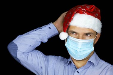Maskeli adam portresi ve siyah arka planda Noel Baba şapkası. Noel kutlaması, Coronavirus salgını sırasında yeni yıl tatili