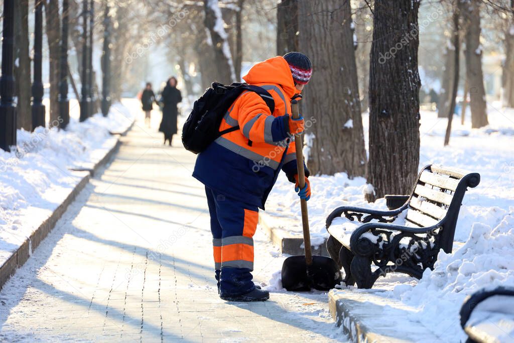 Communal services worker in uniform with a shovel clears melting snow on a sidewalk. Woman during snow removal in winter city, street cleaning