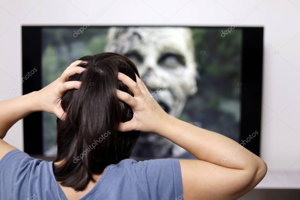 Woman clutching his head looking at zombie on TV screen. Concept of horror movie, fear, scary video