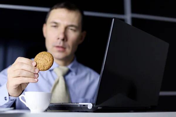 ノートパソコンに座ってオートミールクッキーを食べる男 快適な食べ物 ストレスに対する甘いデザート オフィスでの仕事中の休憩 ストック写真
