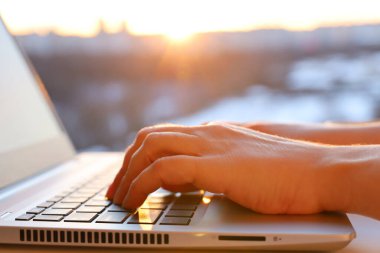 Pencere kenarındaki dizüstü bilgisayarda güneş ışığı gören kadın eller. Konforlu iş yeri, çevrimiçi iletişim, ofis ya da ev işi kavramı