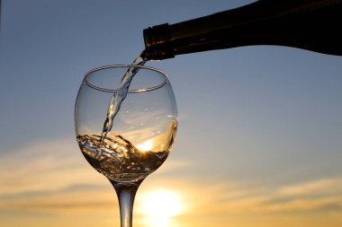 Beyaz şarap, günbatımının arka planında bardağa dökülüyor. Seçici bir odak noktası. Kutlama anlayışı, romantik akşam yemeği.