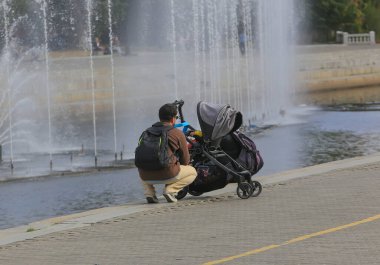 Bebek arabasında bir bebekle Avrupalı baba meydanda bir fıskiyeyle geziniyor.