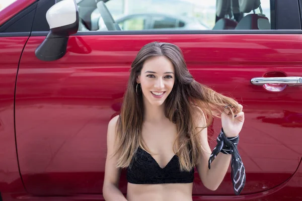 Positive young female in trendy outfit smiling and looking at camera while standing near red car