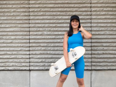 Mavi aktif giysili, siyah şapkalı, kaykaylı uzun siyah saçlı, iyimser genç bir kadın hafta sonu şehir caddesinde sert duvarın yanında durup gülüyor ve uzun siyah saçları düzeltiyordu.