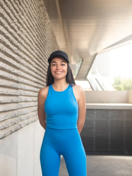Happy young Hispanic woman in blue sportswear and black cap holding hands behind back and looking at camera with smile while standing outside modern building on sunny day