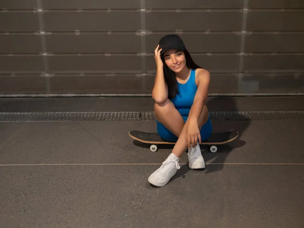 身穿运动服 头戴帽子 快乐的西班牙年轻女子的高视角 在靠近金属墙的时候 坐在滑板上 微笑着看着相机 触摸着头 — 图库照片